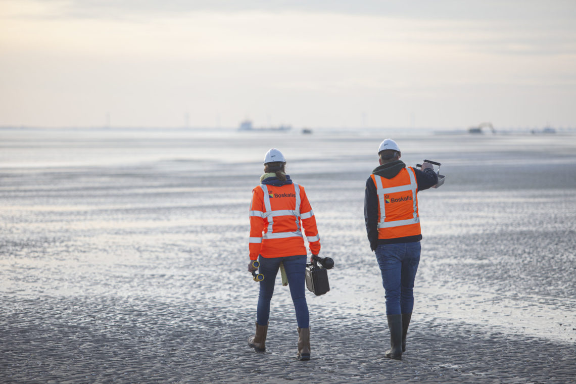 Twee medewerkers van Boskalis lopen over de Roggenplaat in de Oosterschelde. Op de achtergrond zijn kranen aan het werk