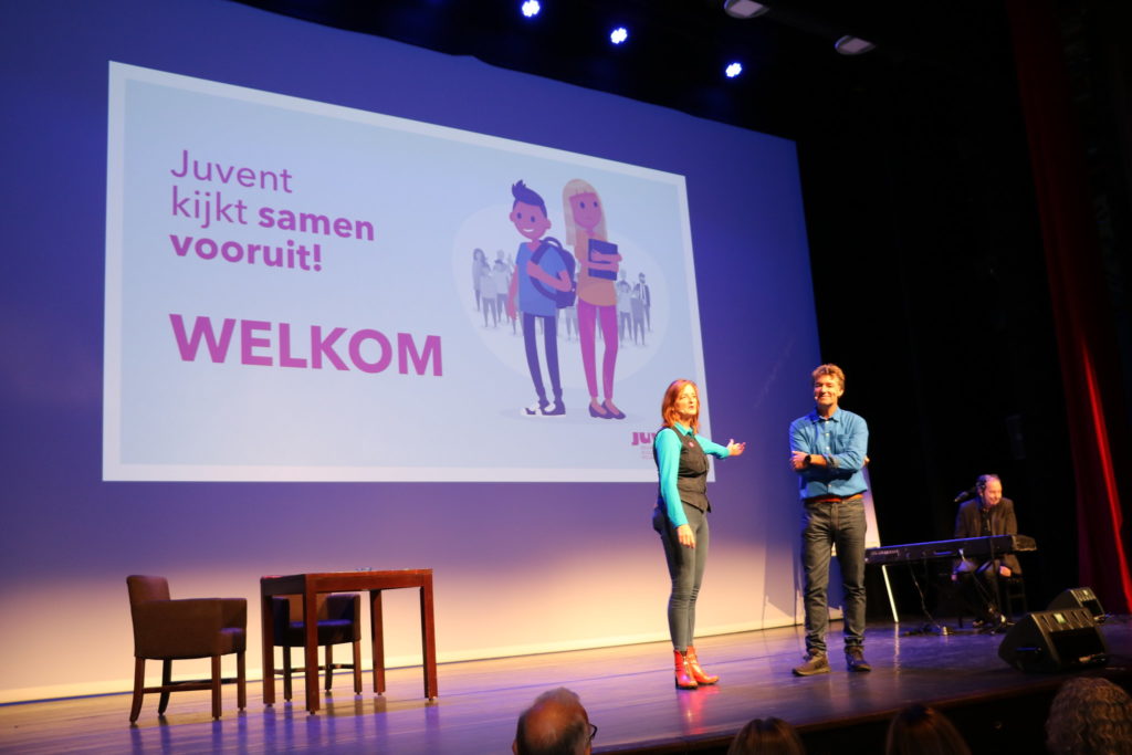 Twee sprekers op een podium verwelkomen de aanwezigen