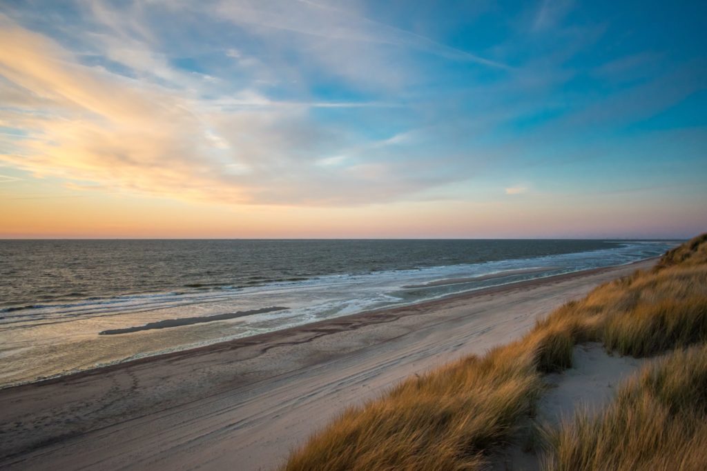 Foto van een ondergaande zon met op de voorgrond de duinen, het strand en de zee