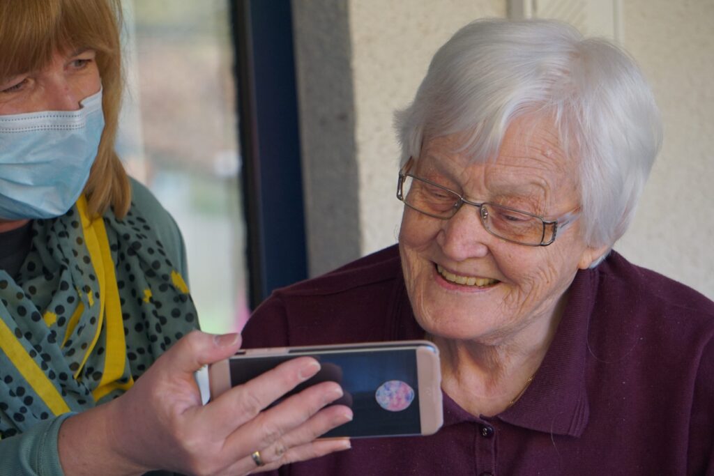 Een oudere mevrouw kijkt op de smartphone van een andere mevrouw. Eén mevrouw heeft een mondkapje op