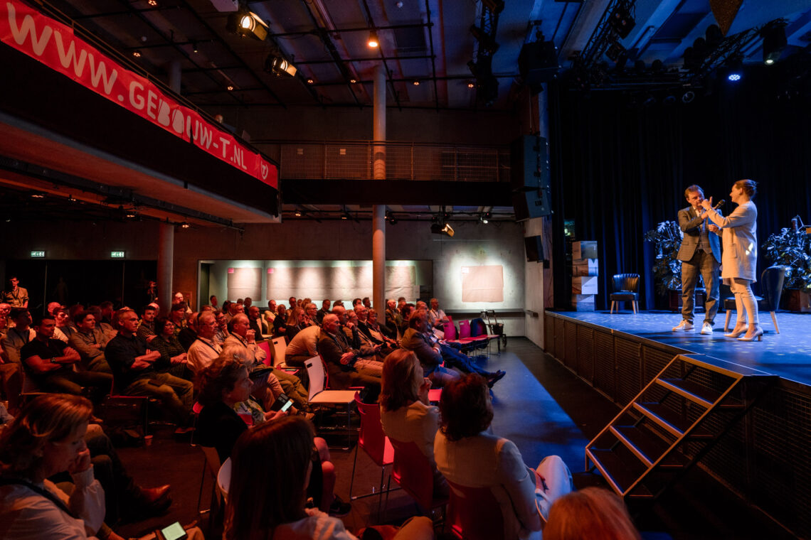 Een man en een vrouw staan op een podium. De zaal is gevuld met publiek.