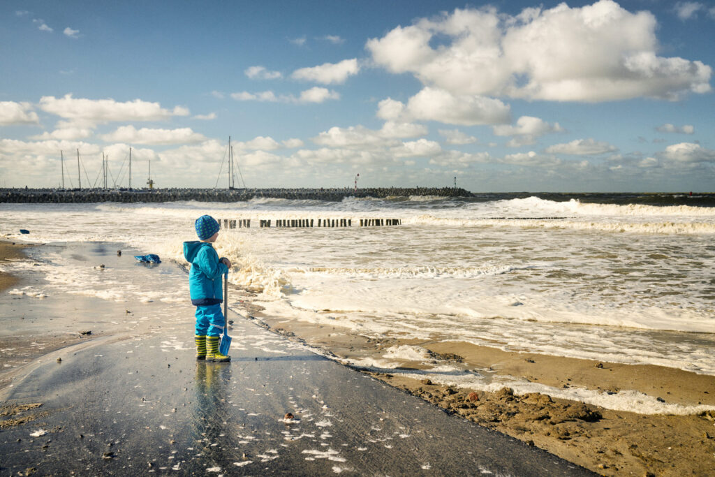 Een kind staat op het strand met een blauw regenpak en muts en gele regenlaarzen, met een schep in zijn hand en kijkend naar de zee. ZWD Cadzand © Kentin Photography