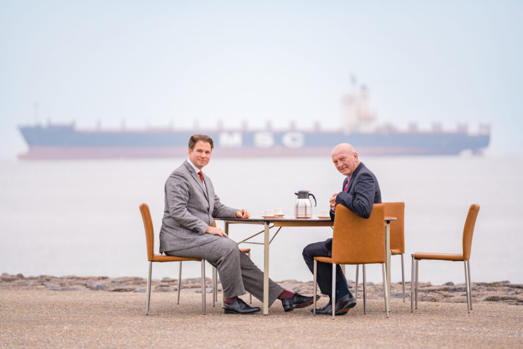 Twee mannen zitten aan een tafel bij de zee koffie te drinken, op de achtergrond staat een groot vrachtschip. Zeeuwse Watertafel © Waterschap Scheldestromen