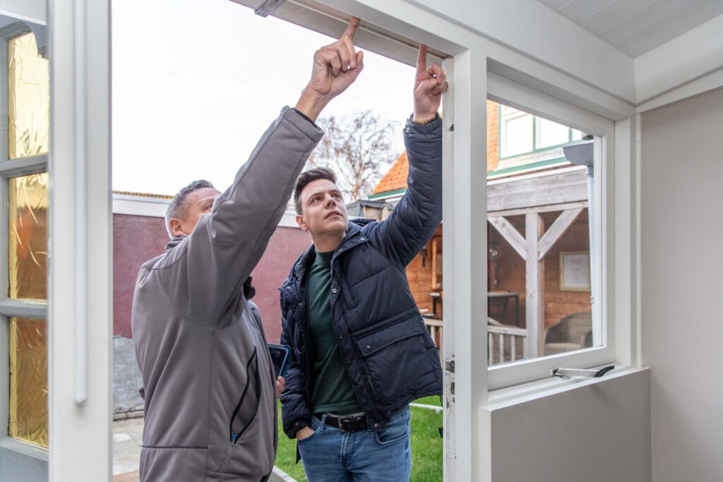 Twee mannen in de deuropening van een huis, ze kijken en wijzen naar het kozijn.