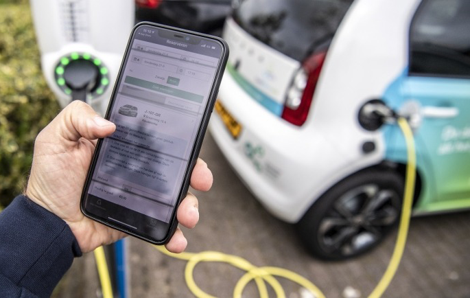 Telefoon in hand bij een laadpaal voor een elektrische auto. De auto is aangesloten aan de lader. Op de telefoon zie je een app met informatie over het opladen.