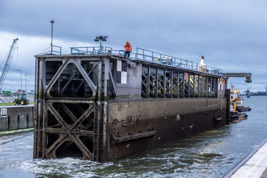 een sluisdeur wordt ingevaren. Hij zit vast an een touw en ligt in het water. bovenop de sluisdeur staat een man met oranje hesje aanwijzingen te geven. Nieuwe Sluis Terneuzen, Lievens Communicatie.