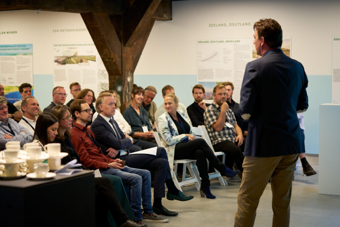 Een man spreekt een volle zaal toe. De mensen luisteren aandachtig