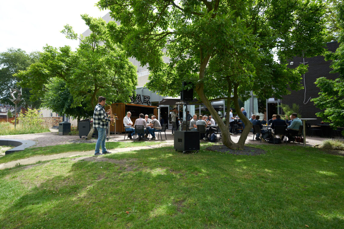 Mensen zitten buiten in de zon op terras, een man speelt Saxofoon.