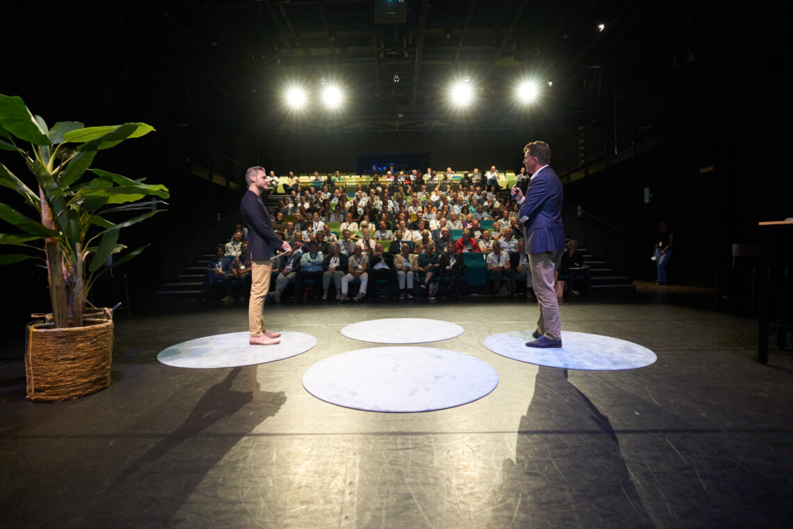 Twee sprekers op het podium spreken een volle zaal toe tijdens de gebiedsconferentie Zuidwestelijke Delta