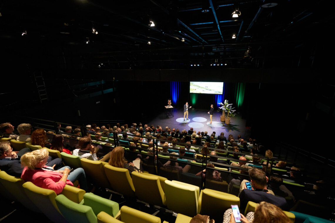 Mensen in een theater luisteren naar twee sprekers op het podium tijdens een conferentie van Zuidwestelijke Delta