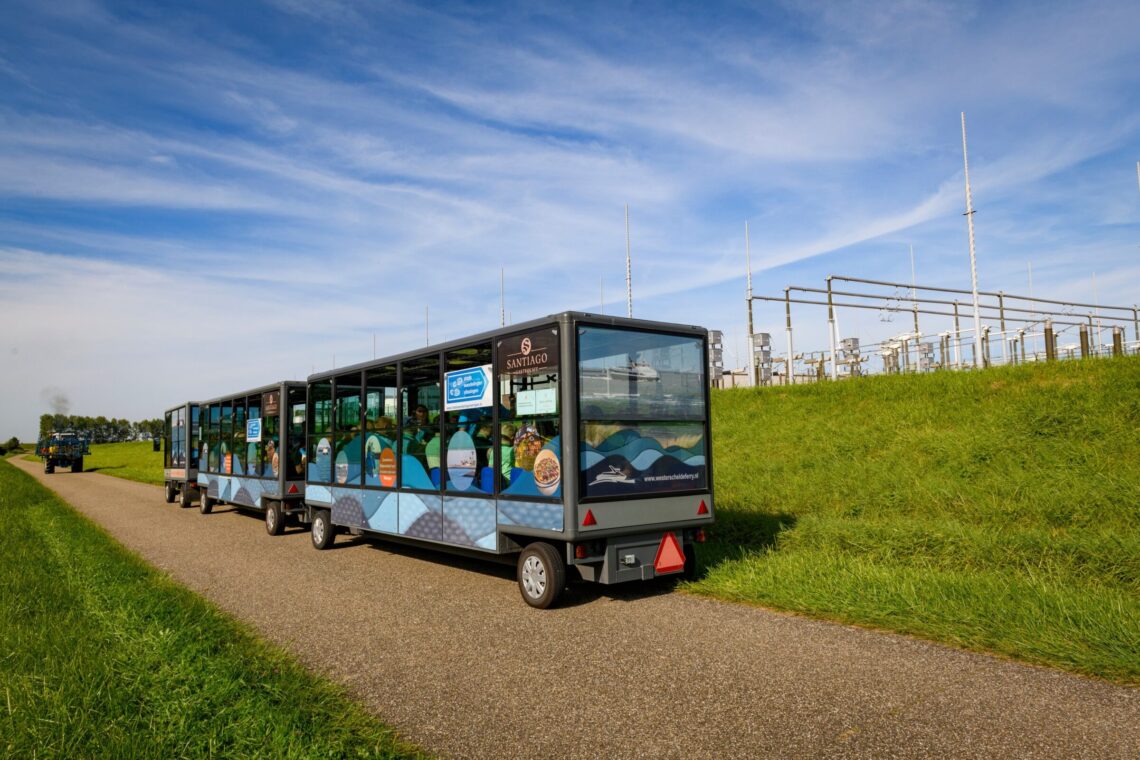 Een elektrische tram rijdt over een weg in de polder langs een hoogspanningsstation. Ervoor rijdt een trekker